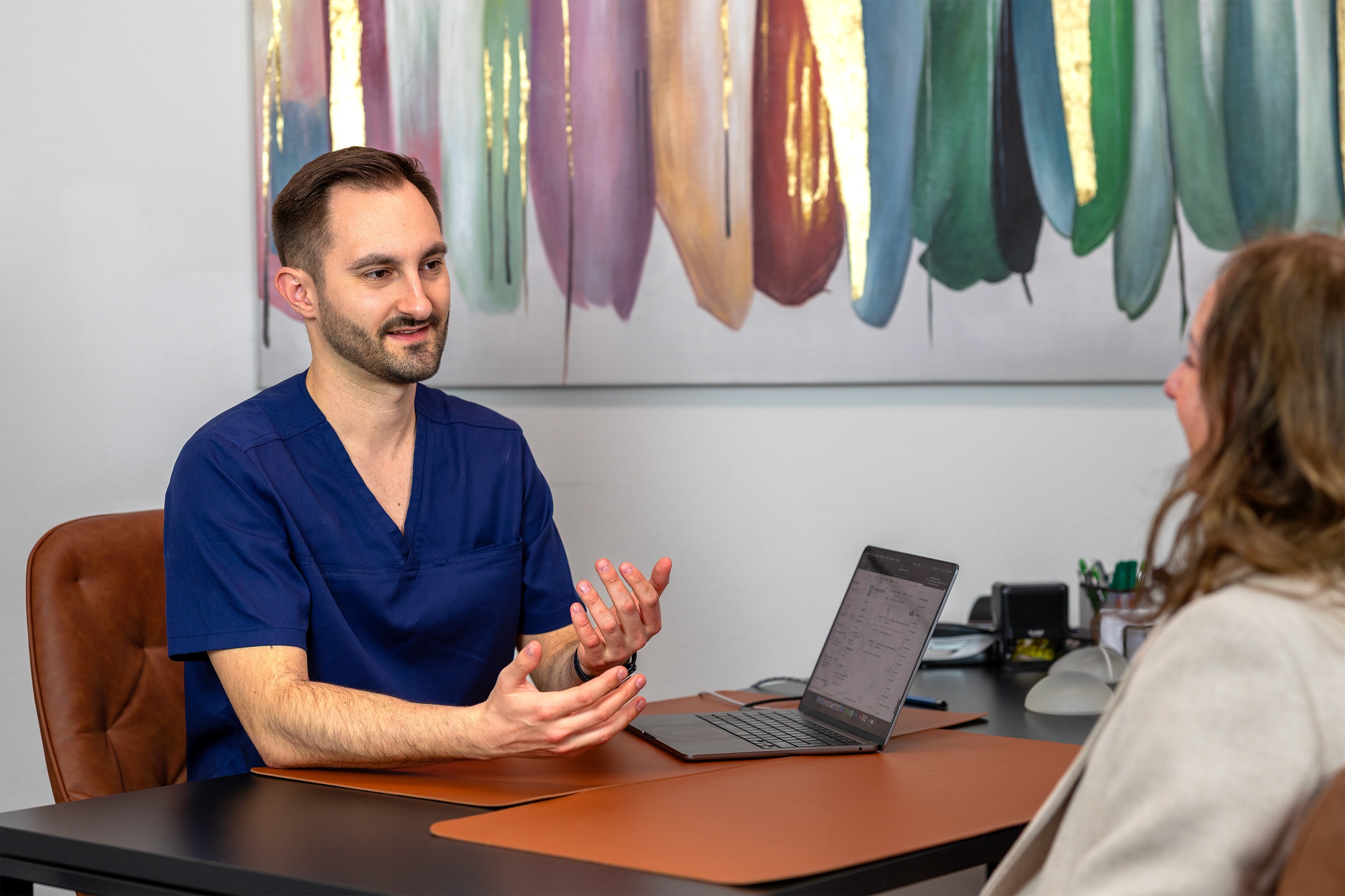 213A0642-2 - Dr. Pumberger Medical professional in blue scrubs explains something to a patient across a desk, with a laptop open between them in a clinical office.