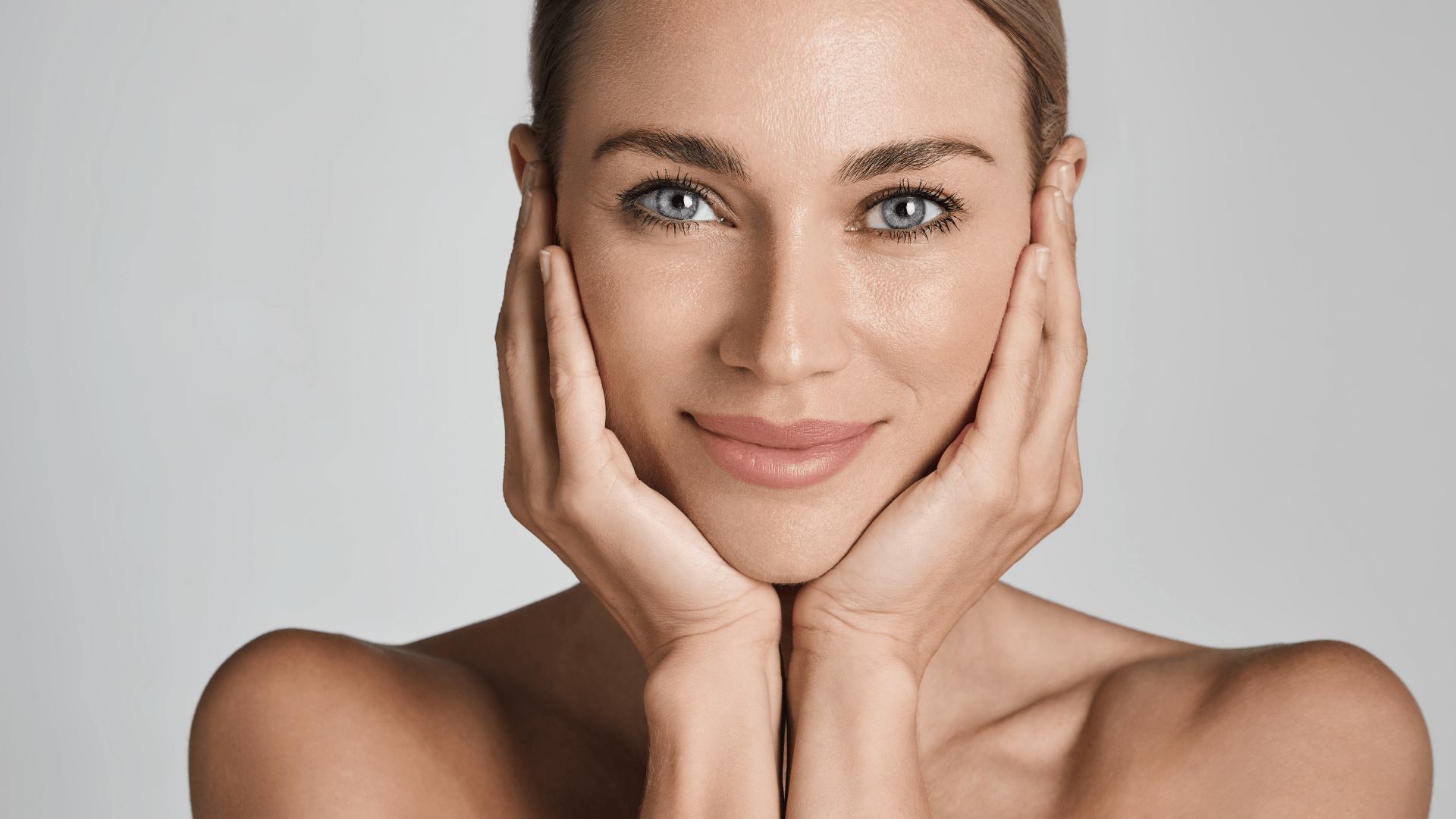Close-up of a smiling woman cupping her face with both hands against a neutral background, portrait orientation