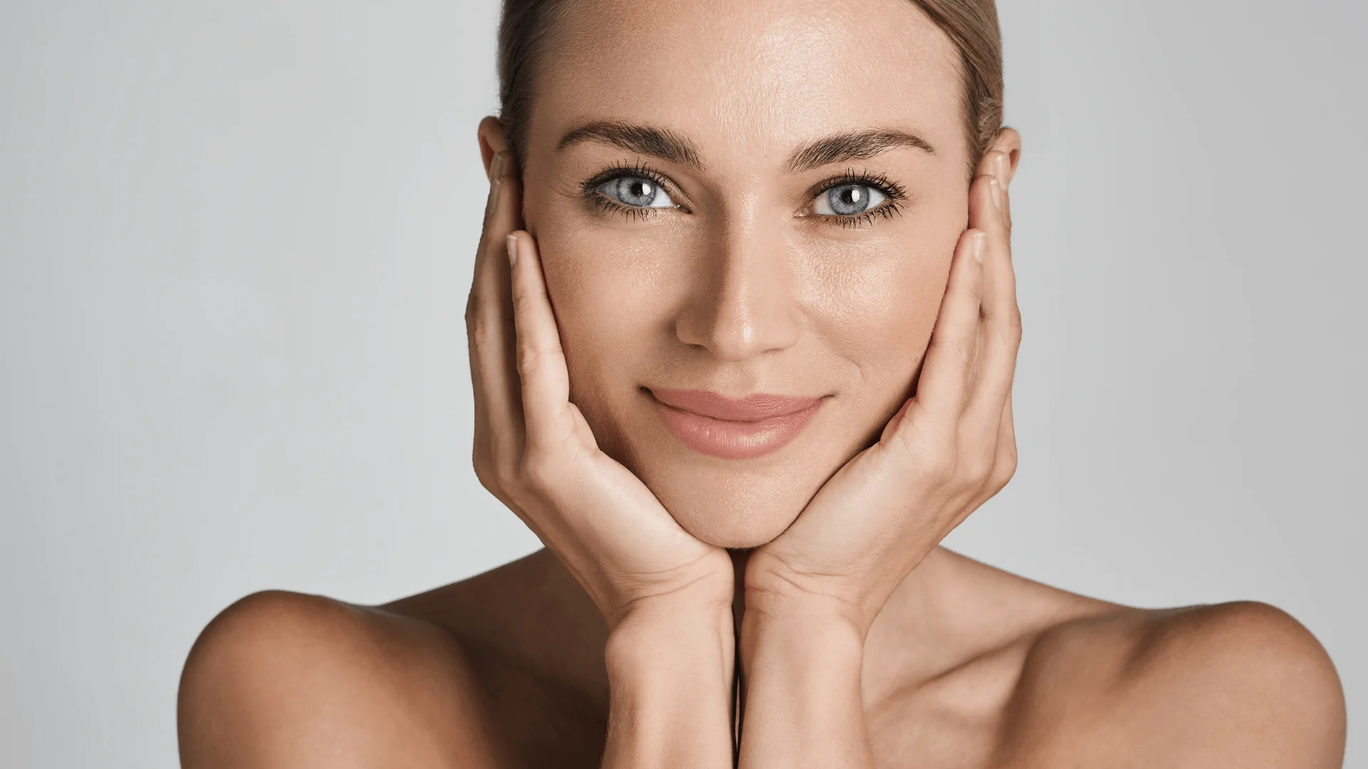 Close-up of a smiling woman cupping her face with both hands against a neutral background, portrait orientation