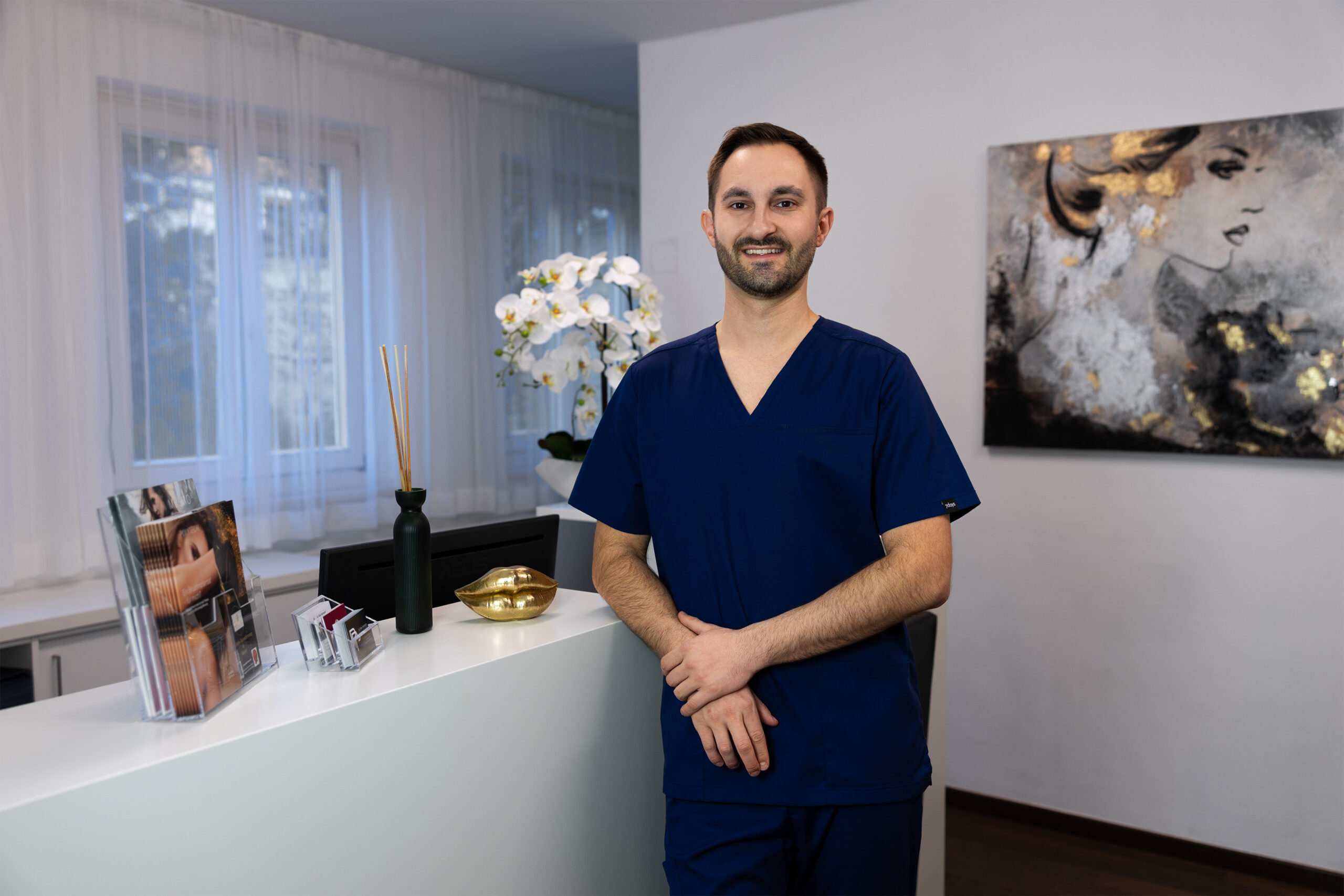 Website 14 - Dr. Peter Pumberger Smiling male healthcare worker in blue scrubs stands at a reception desk in a bright clinic.