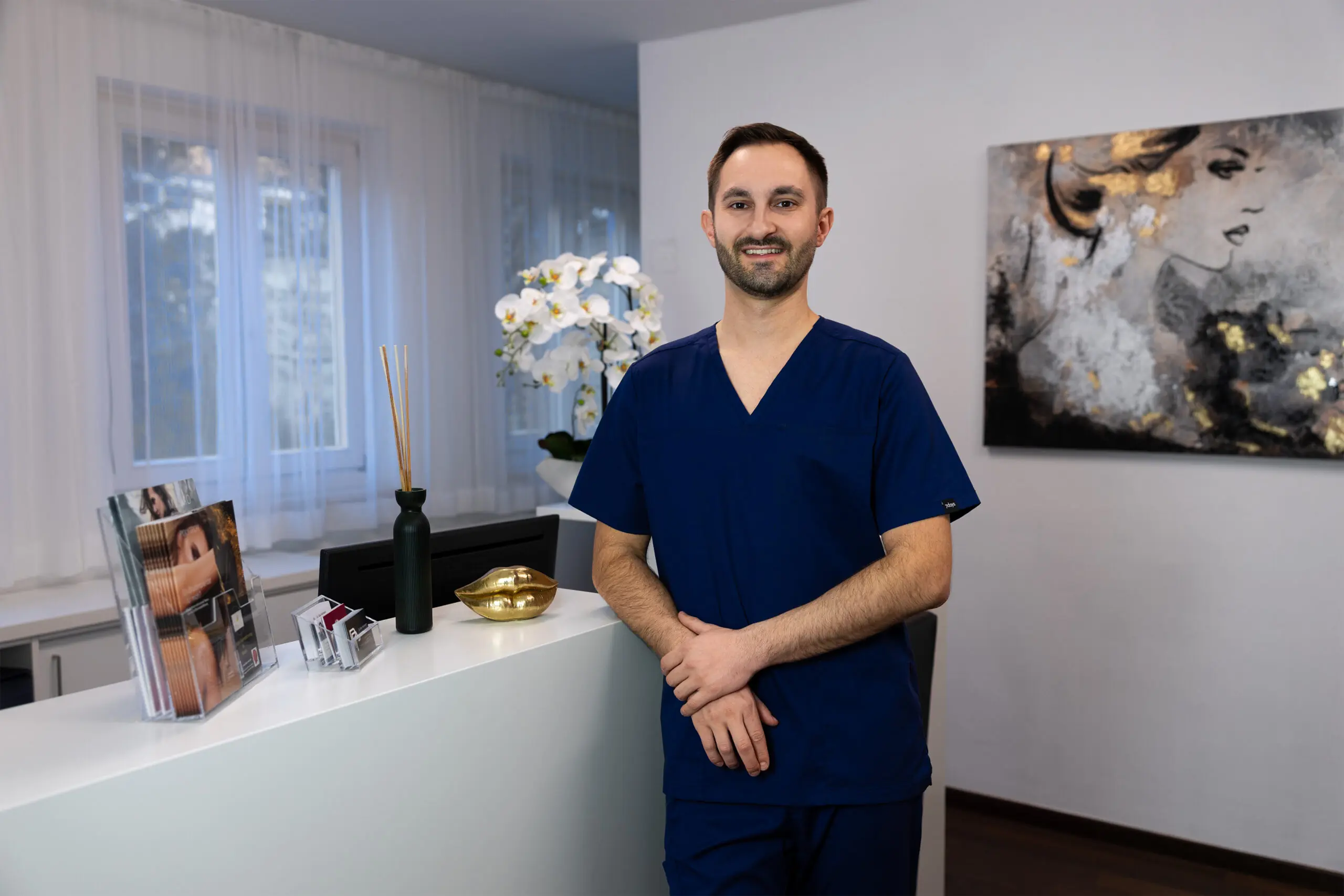 Website 14 - Dr. Pumberger Smiling male healthcare worker in blue scrubs stands at a reception desk in a bright clinic.